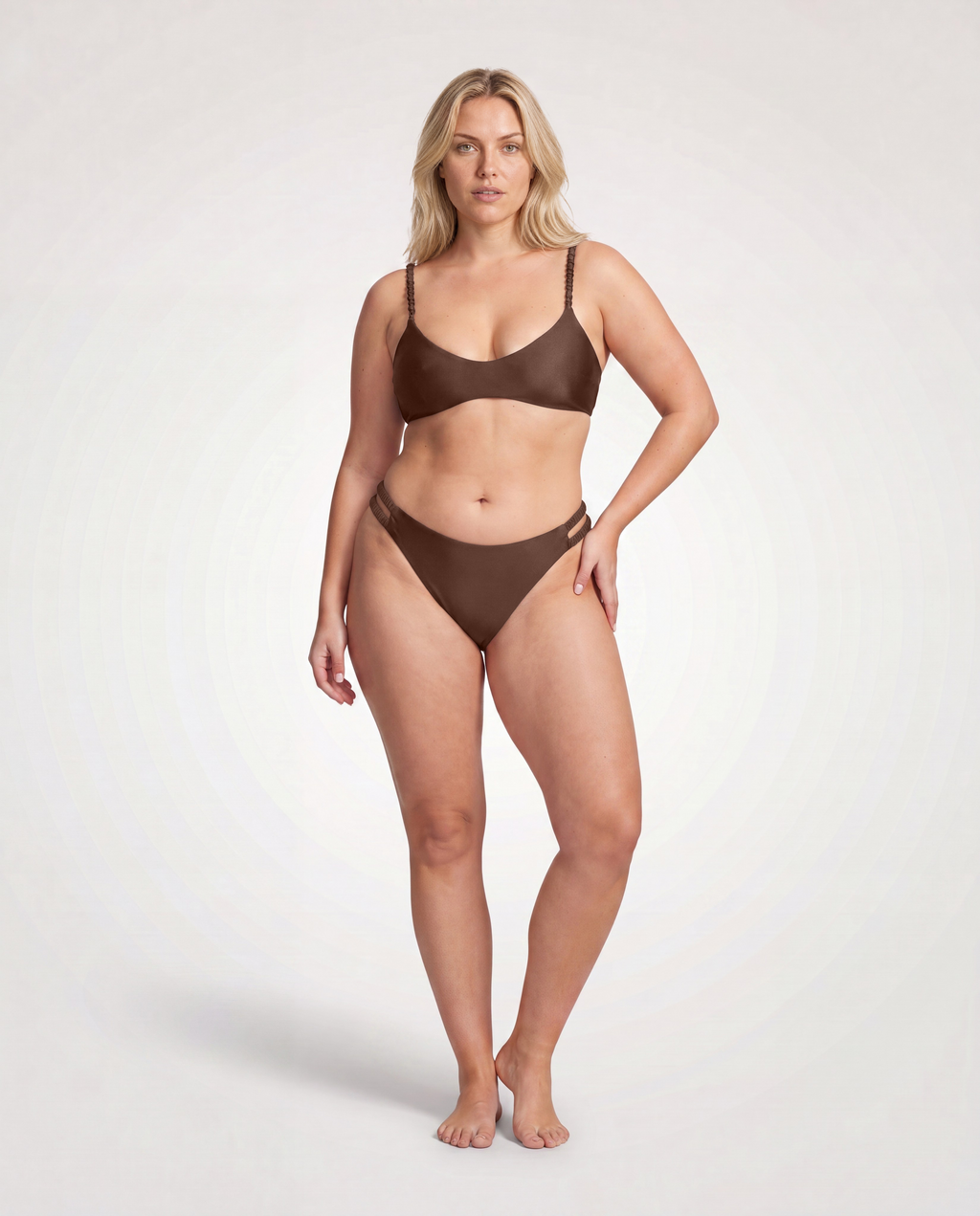 A blonde model stands against a light gray studio background wearing a matching two-piece bikini in a dark brown color, featuring a scoop-neck triangle top and matching high-cut bottoms.